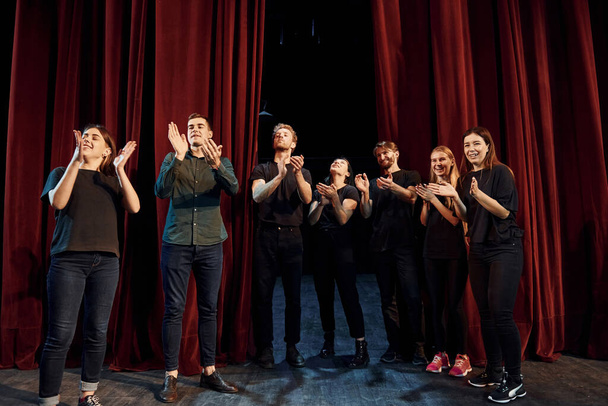 Standing against red curtains. Group of actors in dark colored clothes on rehearsal in the theater. - Foto, imagen
