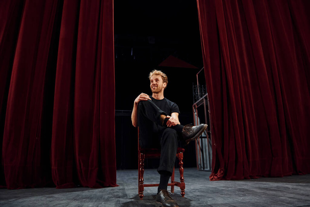 Positive young man sitting on the chair between the curtains on the stage of theater. - Foto, Bild