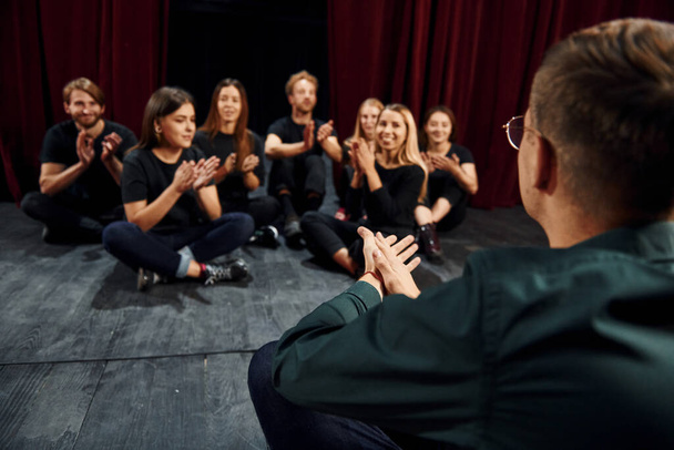 Op de grond zitten. Groep acteurs in donker gekleurde kleren op repetitie in het theater. - Foto, afbeelding