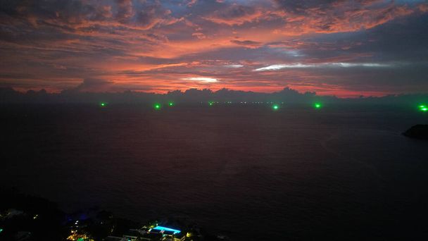 Top view of the red sunset, ocean and clouds. The sky is reflected in the water of the sea. Green lights are burning. Palm trees grow on island. Roofs of hotels and roads are visible. Boat is sailing - Fotoğraf, Görsel