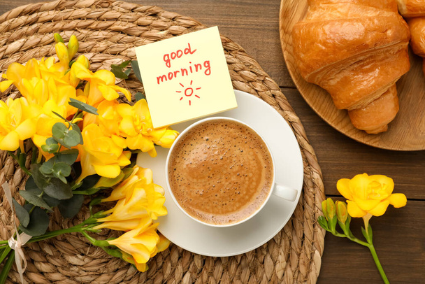 Cup of aromatic coffee with croissant, beautiful yellow freesias and Good Morning note on wooden table, flat lay - Photo, Image