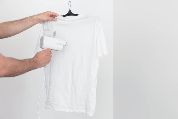 Cocine a vapor la camiseta con un vapor portátil sobre un fondo blanco. Primer plano. espacio libre para texto - Foto, imagen