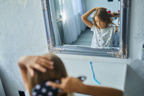 Pretty Little girl peinándose sola el pelo delante de un espejo, cepillándose el pelo en casa, niño sonriendo, rutina de la mañana - Foto, imagen