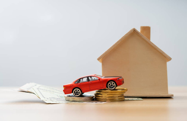 Rode miniatuur auto model op een stapel munten in de voorkant van een houten huis naast een bankbiljet, Concept van Auto leningen en woningverzekering, economische problemen en inflatie - Foto, afbeelding