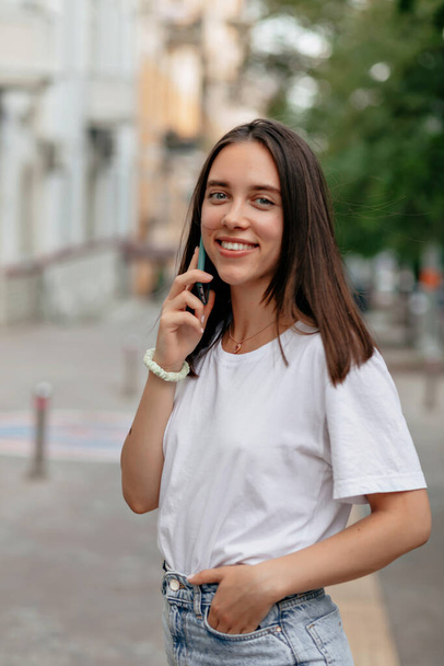 Close-up portret van schattige mooie vrouw met vrolijke glimlach en donker haar draagt witte t-shirt en jeans is praten op de telefoon en glimlachen op wazige stad achtergrond - Foto, afbeelding