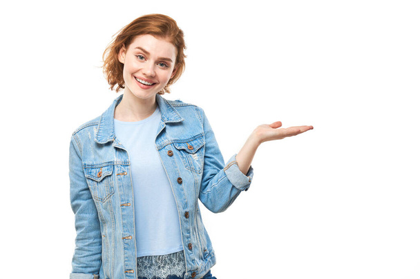 Attraente rossa promoter ragazza in camicia di jeans che tiene qualcosa in mano dimostrando spazio vuoto con viso eccitato isolato su sfondo bianco - Foto, immagini