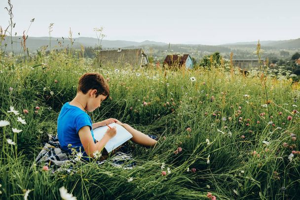 The boy sits in the grass on the mountain and draws a picture on the top of the mountain. A young artist paints a mountain landscape in summer. - Φωτογραφία, εικόνα