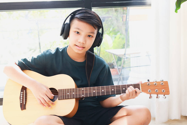 Asiatico ragazzo indossa cuffie con musica nera Sedetevi e suonare la chitarra in casa. Concetto di apprendimento musicale, formazione sulla competenza musicale. - Foto, immagini