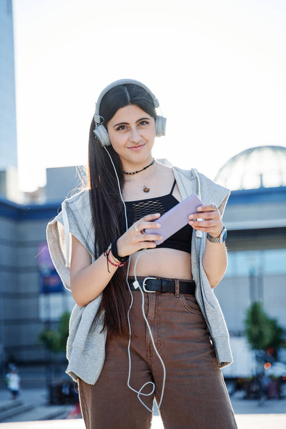 スマートフォンを屋外で、ヘッドフォンを身に着けている若い女性 - 写真・画像