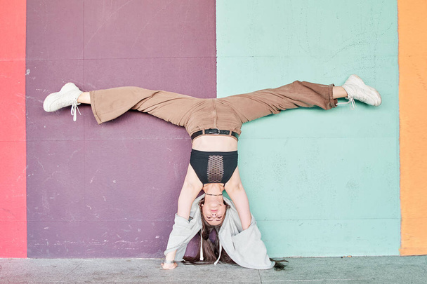 Full length powerful woman in casual wear performing handstand with split near colorful wall - Foto, Imagen