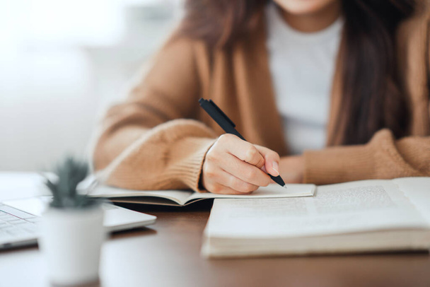 Close-up - Hand van de vrouw schrijven in spiraal notitieblok geplaatst op houten bureaublad met verschillende items thuis. - Foto, afbeelding