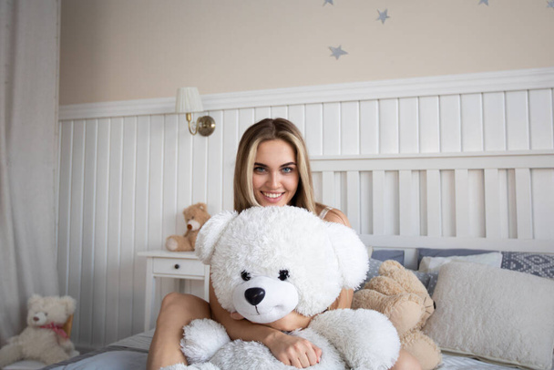 Cute blonde woman sitting with teddy bear on bed in bedroom. - Фото, изображение