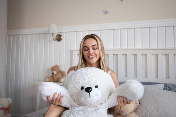 Cute blonde woman sitting with teddy bear on bed in bedroom. - Fotoğraf, Görsel