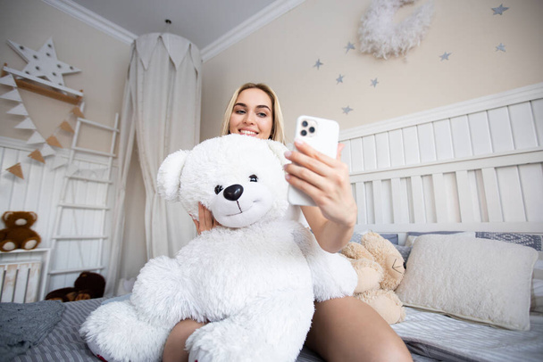 Hermosa mujer rubia hace una selfie con un oso de peluche sentado en la cama. - Foto, imagen