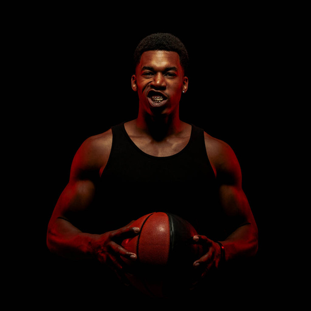 Basketball player side lit with red color holding a ball against black background. African american man silhouette with face gesture. - Foto, Bild