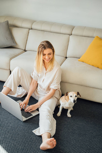 Beautiful blonde woman using laptop. working from home, studing or online shopping. Freelancer or influencer remote job. Sitting on floor in living room with small cute dog. Vertical composition - Photo, Image