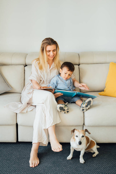 Family reading a book. Woman her son and small dog in living room reading book sitting on a beige sofa in sunny living room. early development of child. reading fairy tales and children's literature - 写真・画像