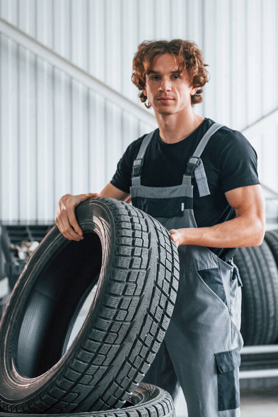 Holds tire. Adult man in grey colored uniform works in the automobile salon. - Photo, Image