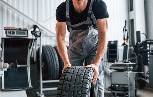 Rolls the tire. Adult man in grey colored uniform works in the automobile salon. - Фото, зображення