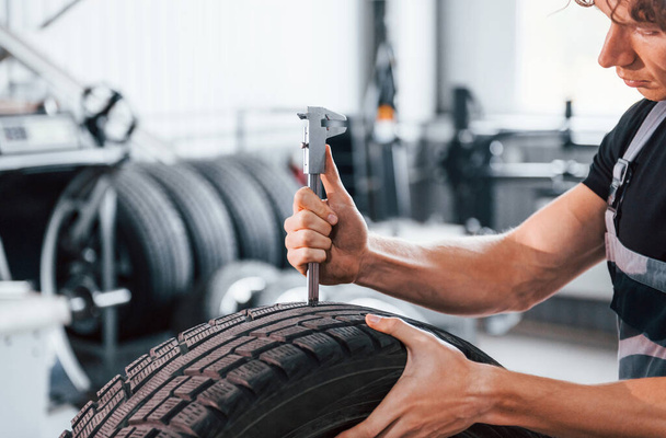 Works with tire. Adult man in grey colored uniform is in the automobile salon. - 写真・画像