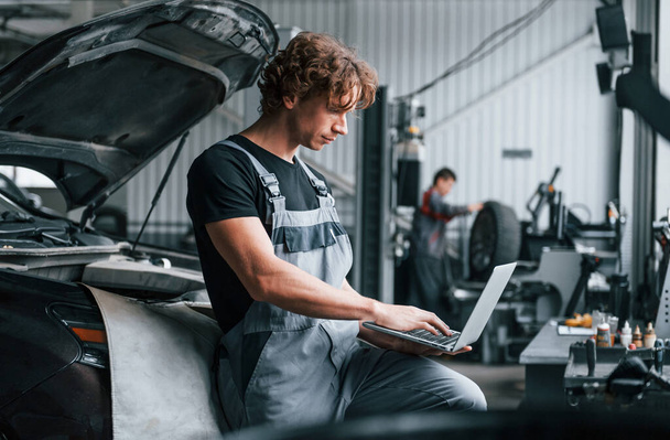 With laptop. Adult man in grey colored uniform works in the automobile salon. - Foto, imagen