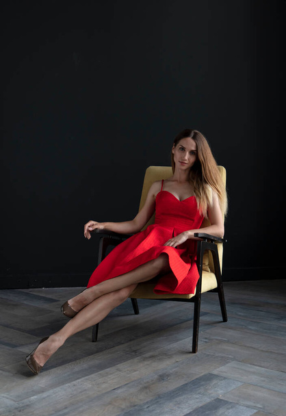 portrait of beautiful young woman sitting on chair in red dress on dark background - Photo, Image
