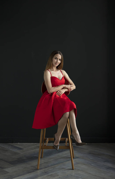 portret van mooie jonge vrouw zittend op stoel in rode jurk op donkere achtergrond - Foto, afbeelding