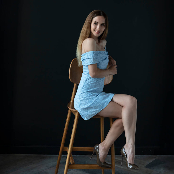 portrait of beautiful young woman sitting on chair in blue dress on dark background - Zdjęcie, obraz