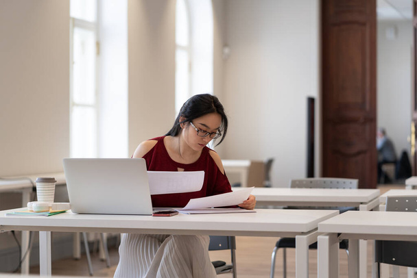 Азійська дівчина студент коледжу в окулярах читає матеріали під час підготовки до іспиту або іспиту в бібліотеці - Фото, зображення