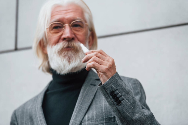 Senior businessman in formal clothes, with grey hair and beard is outdoors smokes electronic cigarette. - Foto, Imagen