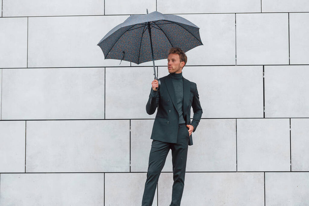 Young man in formal clothes with umbrella standing near wall outdoors with umbrella. - Фото, изображение