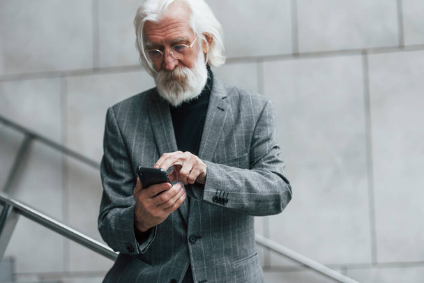 Senior businessman in formal clothes, with grey hair and beard is outdoors trying wireless headphones with phone. - Foto, immagini