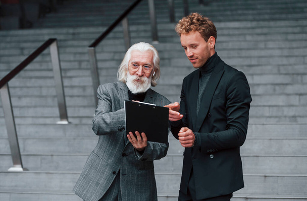 Reading plan. Young guy with senior man in elegant clothes is outdoors together. Conception of business. - Foto, Imagen