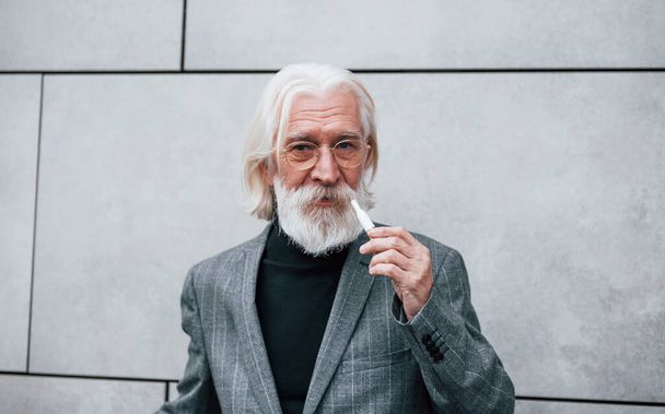 Empresário sênior em roupas formais, com cabelos grisalhos e barba é ao ar livre fuma cigarro eletrônico. - Foto, Imagem