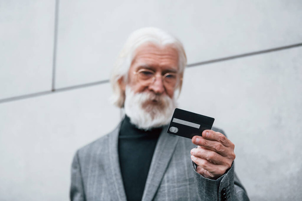 Senior businessman in formal clothes, with grey hair and beard is outdoors holds credit card. - Fotoğraf, Görsel