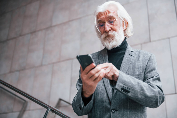 Using smartphone. Senior businessman in formal clothes, with grey hair and beard is outdoors. - Фото, изображение