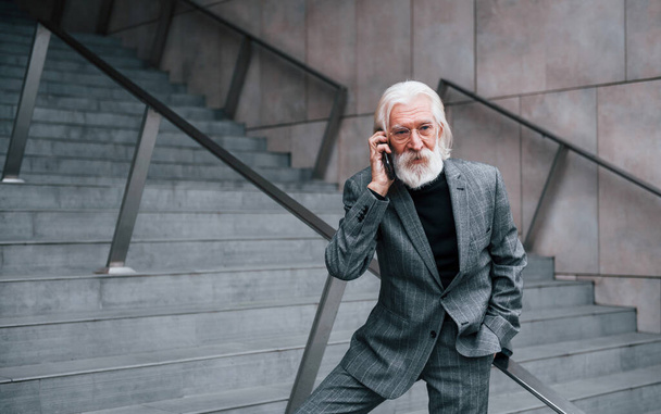 Senior businessman in formal clothes, with grey hair and beard is outdoors talks by phone. - Foto, Imagen