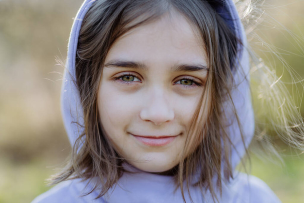 A portrait of pretty child girlin hoodie standing in summer park looking at camera smiling happily. - Фото, зображення