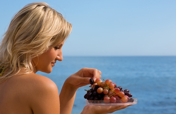 κορίτσι τρώει τα σταφύλια στην παραλία δίπλα στη θάλασσα. - Φωτογραφία, εικόνα