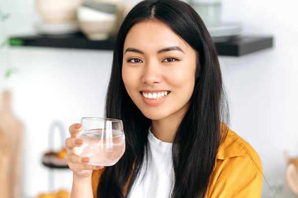 Pretty, healthy chinese brunette girl, dressed in casual stylish clothes, holds a glass of pure water, takes care of her health, drinks daily norm of water, looking at camera, smiles friendly - Valokuva, kuva