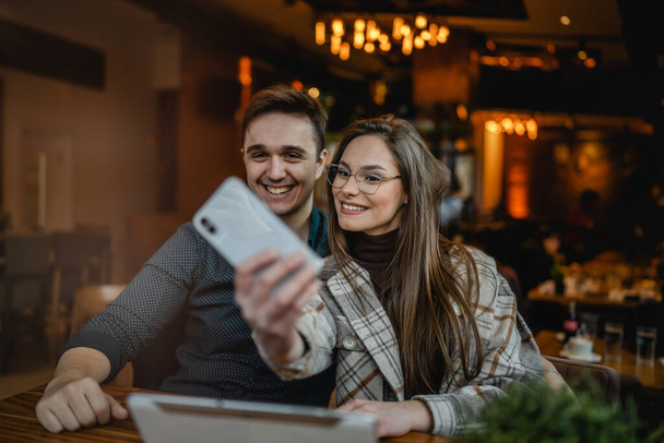 Two people young caucasian couple man and woman female and male friends or boyfriend and girlfriend taking selfie photos or making a video call while sitting at cafe copy space fun and romance - Fotó, kép
