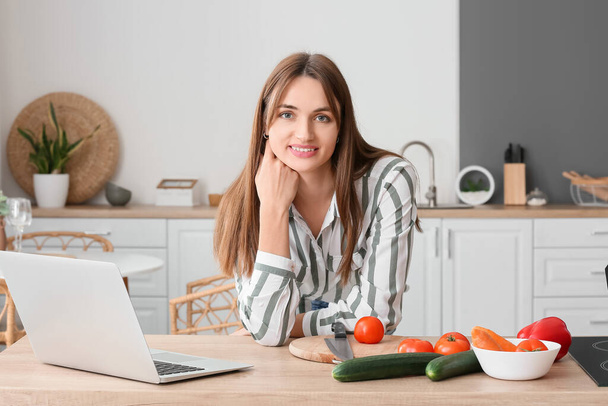 Όμορφη νεαρή γυναίκα με φορητό υπολογιστή και φρέσκα λαχανικά στην κουζίνα - Φωτογραφία, εικόνα