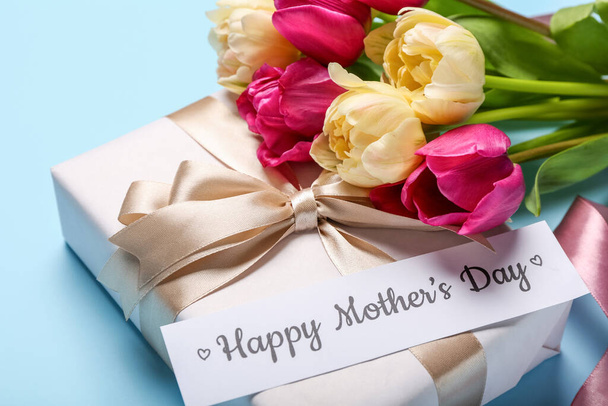 Paper with text HAPPY MOTHER'S DAY, gift box and bouquet of tulips on blue background, closeup - Photo, Image