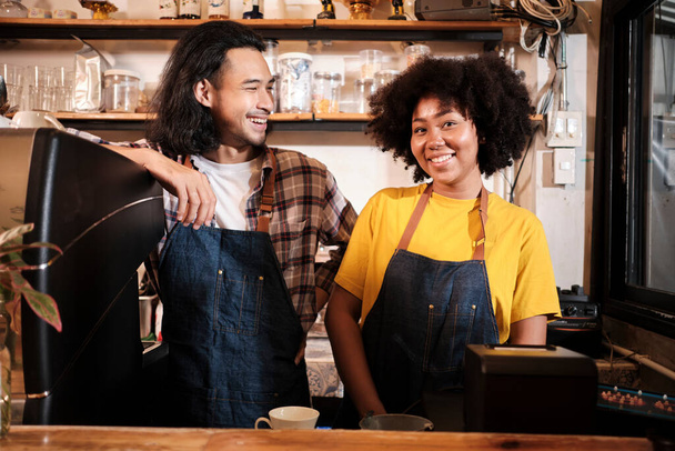 Two cafe business startup partners and friends, African American female, and Thai male baristas talk and cheerful smile together at counter bar of coffee shop, happy service job, and SME entrepreneur. - Foto, immagini