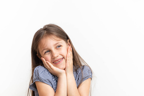 Cute 7 year old girl with long hair smiles, holding hands on face and not looking at camera - Photo, image