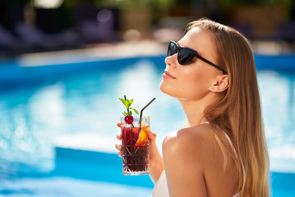 Blonde woman drinks refreshing cocktail sunbathing and sitting near swimming pool at tropical spa. Female in sunglasses enjoys a drink on poolside. Girl chilling in tropical resort on vacation. - Фото, изображение