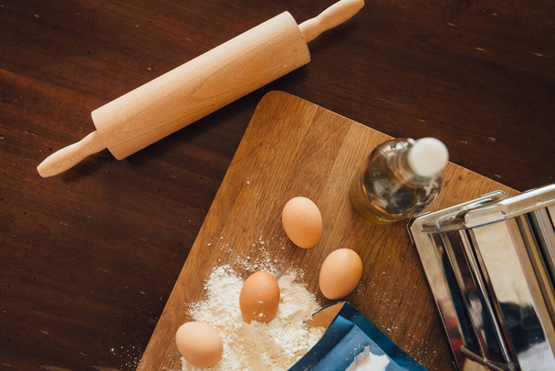 Flour, eggs, olive oil, paste machine on the cutting board ready for homemade italian pasta preparation - Photo, image