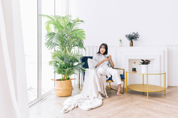 Jonge mooie vrouw glimlachen en zit in de stoel van de moderne woonkamer met een groene plant, Ontspannen het drinken van een kopje hete koffie en genieten van een rustige tijd en een overdekte omgeving in gezellig huis. - Foto, afbeelding