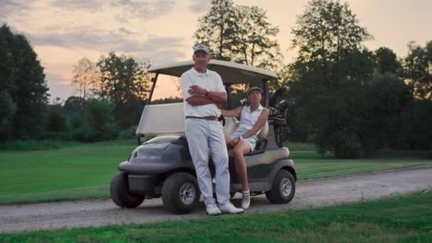 Pareja posando carrito de golf afuera. Dos golfistas toman equipos deportivos clubes en el campo - Metraje, vídeo