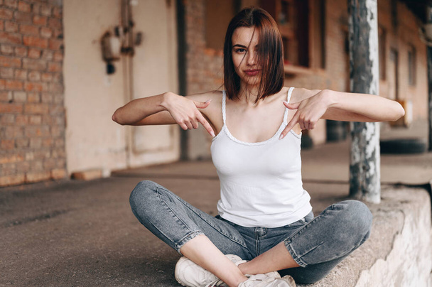 A girl with a short, shoulder-length haircut sits outside and points her index fingers down to indicate what. A vertical photo of the girl sits cross-legged. - Photo, Image